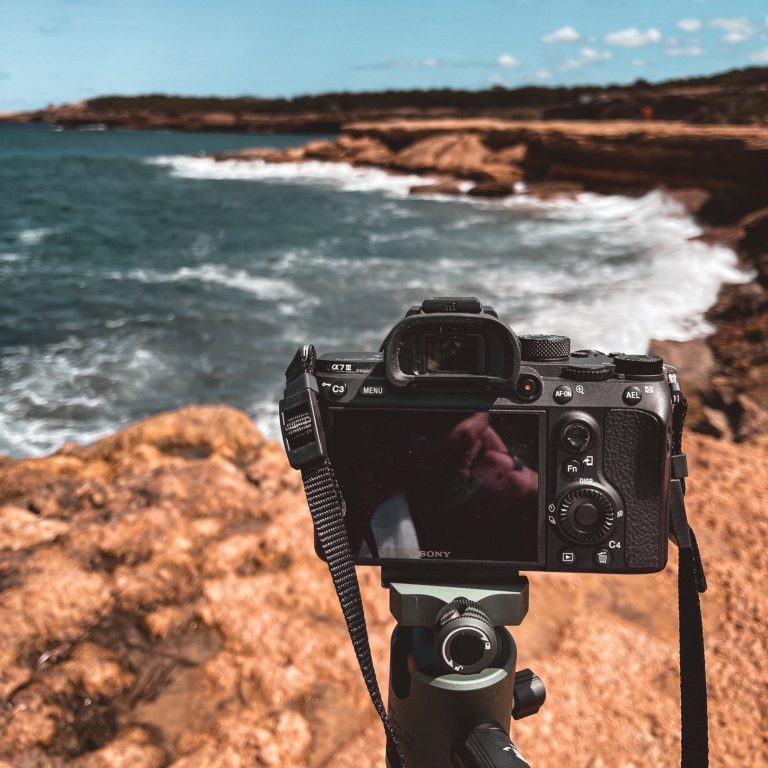 Fotografie oliventure.media Kamera auf einem Stativ am Meer mit Wellen und felsiger Küste im Hintergrund.